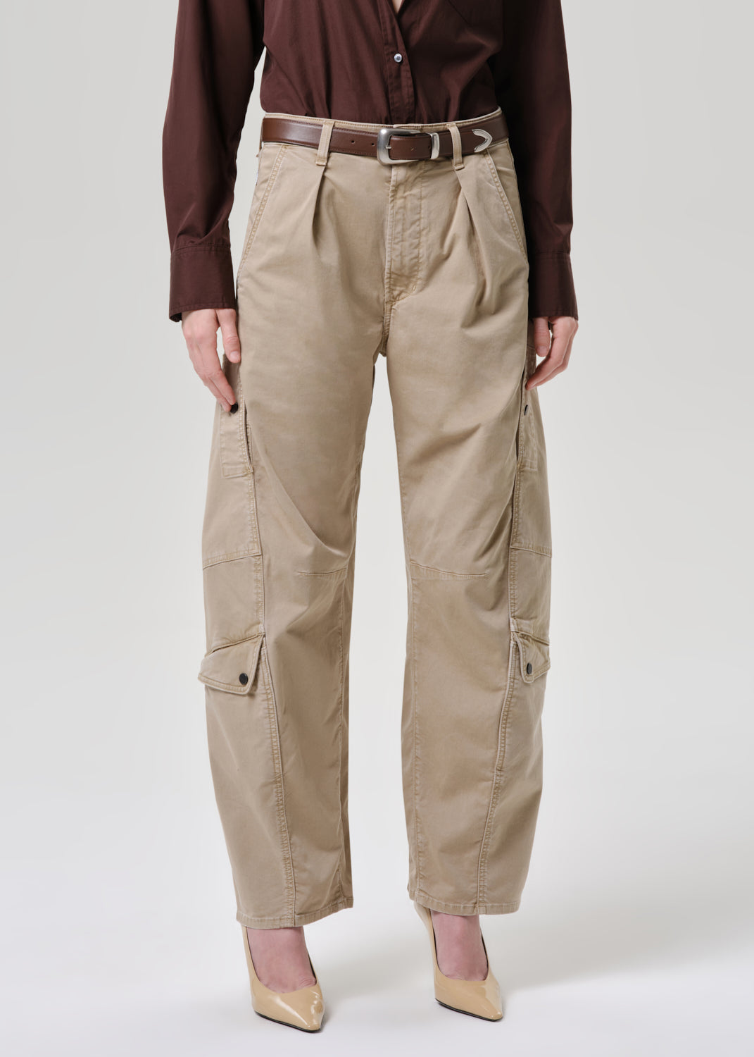 A model is shown from the waist down wearing beige cargo pants with a brown belt, a dark brown button-up shirt, and beige heels against a grey background.