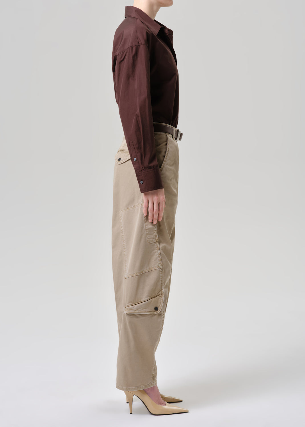 A model shown from the side wearing a dark brown long-sleeve collared shirt tucked into beige cargo pants with a dark belt, paired with beige high-heeled pumps, against a grey background.