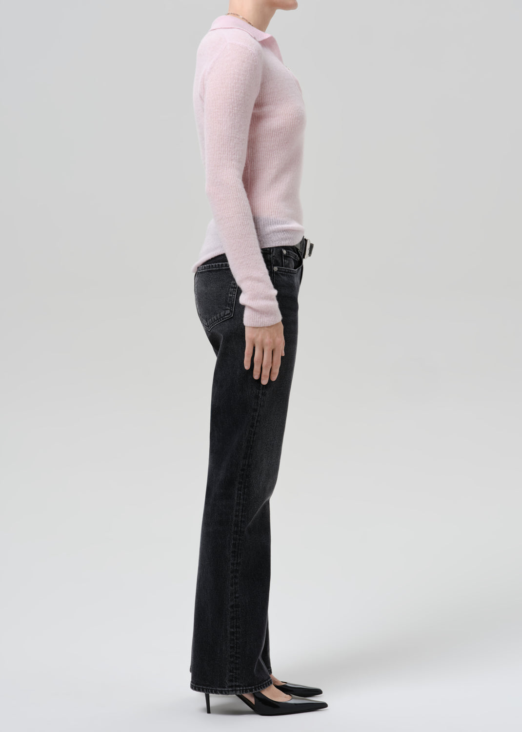 Person wearing a light pink ribbed V-neck sweater with black straight leg jeans viewed from the side on a grey background