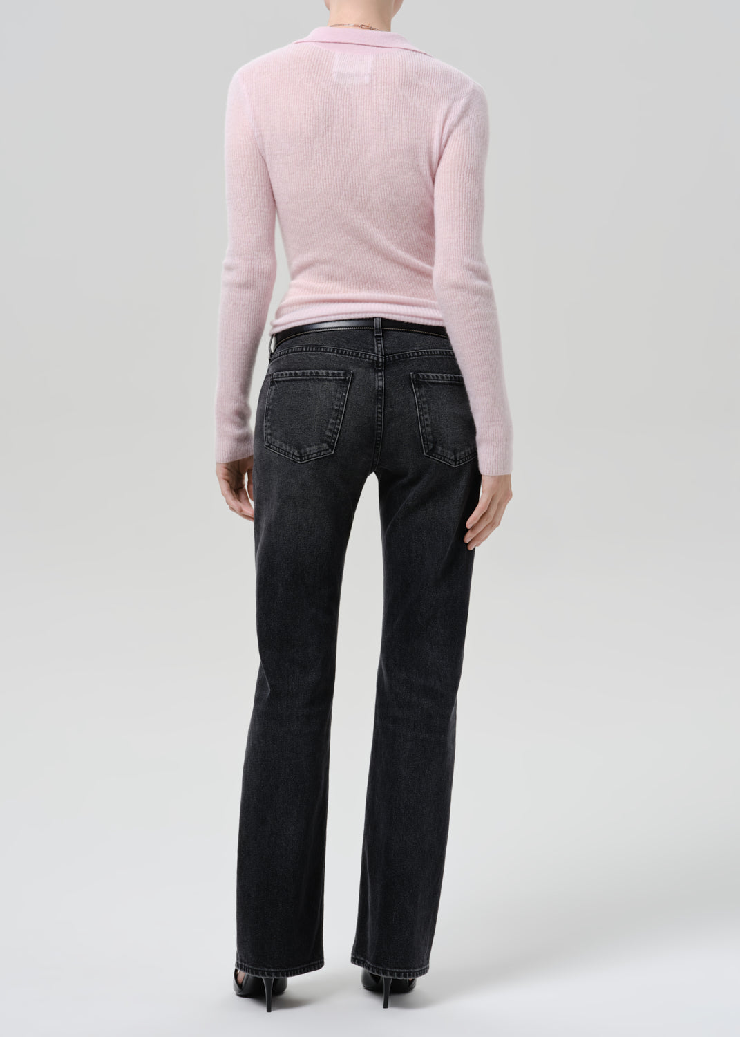Person wearing a light pink ribbed V-neck sweater with black straight leg jeans viewed from the back on a grey background