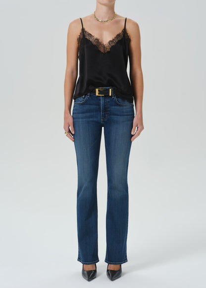A model wearing a black satin camisole top with black lace trim, tucked into blue jeans with a gold belt, standing against a grey background.