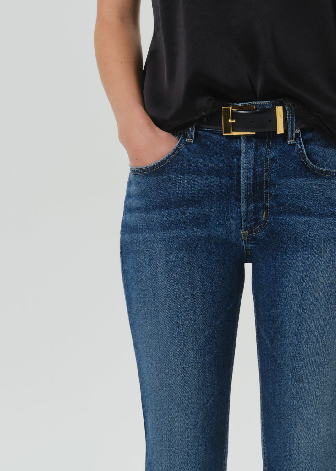 Close-up of a person wearing dark blue denim jeans with a black belt featuring a gold buckle, paired with a black top against a grey background.