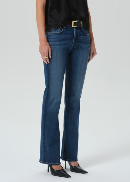 A model wearing a pair of dark blue straight-leg jeans with a gold-tone belt buckle, paired with a black top and black pointed-toe heels, shown from the waist down on a grey background.