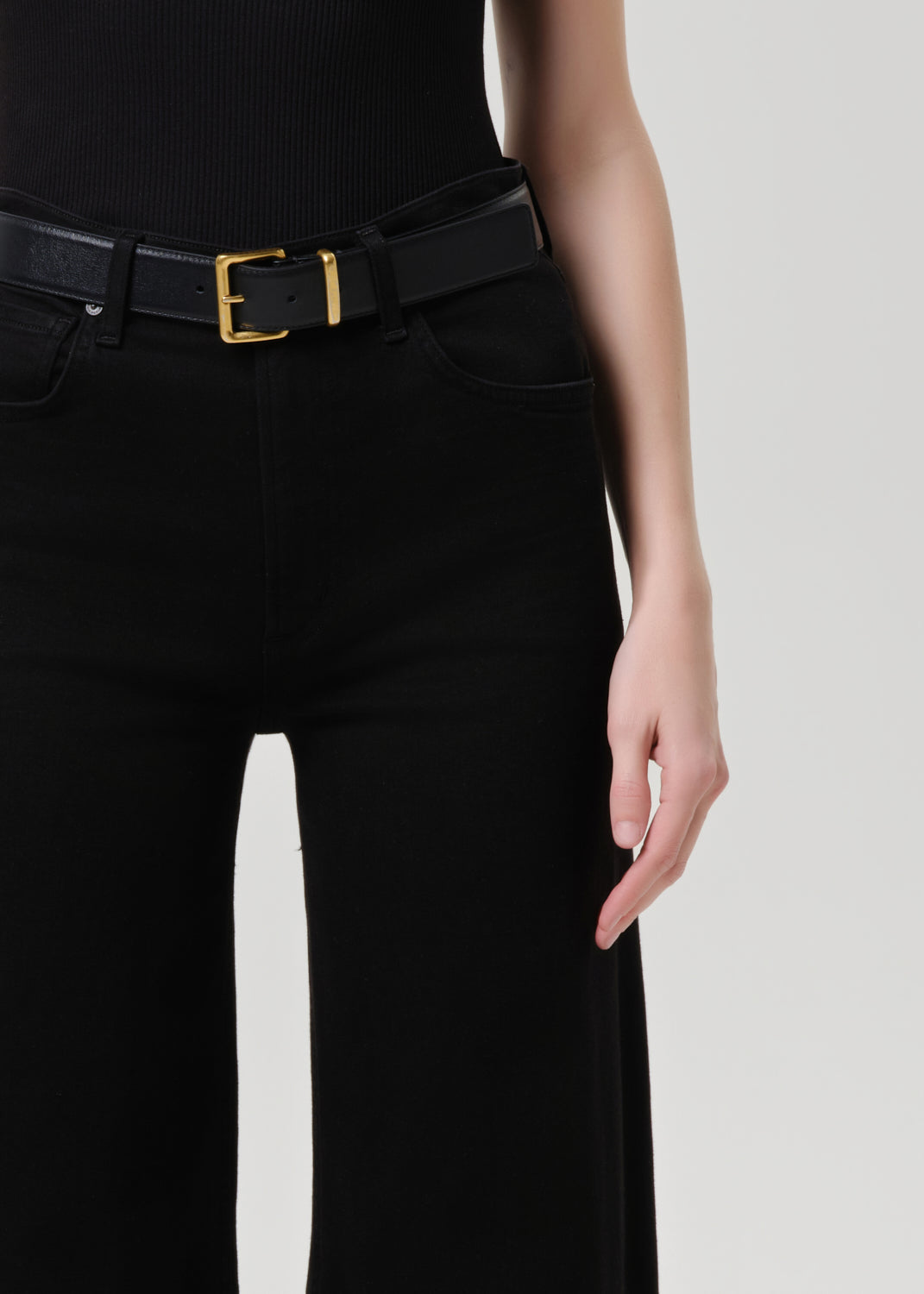 Close-up of a model wearing a black belt with a gold rectangular buckle, paired with black pants and a black ribbed top against a grey background.