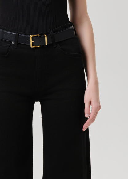 Close-up of a model wearing a black belt with a gold rectangular buckle, paired with black pants and a black ribbed top against a grey background.