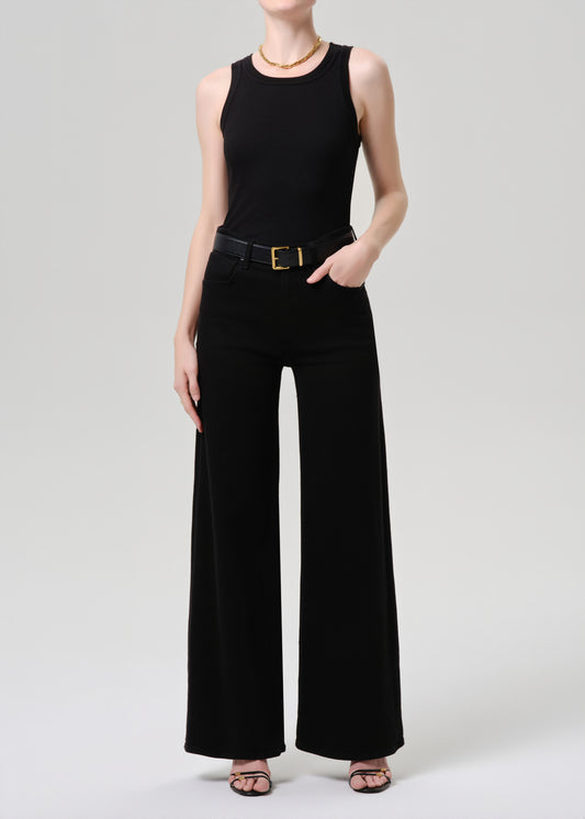 A model wearing a black sleeveless top and matching wide-leg trousers with a black belt featuring a gold buckle, photographed against a grey background.