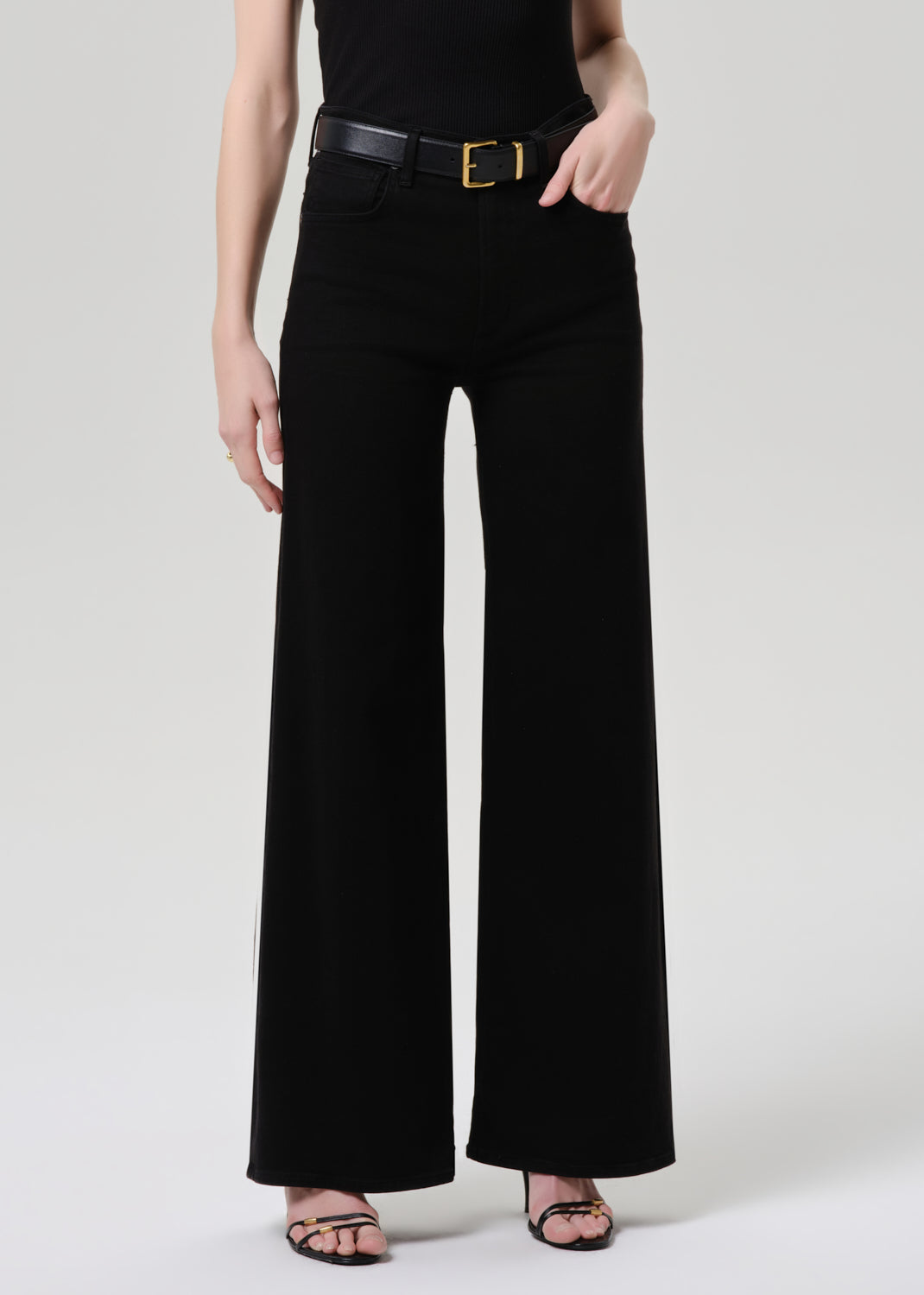 A model wearing black wide-leg trousers with a black belt featuring a gold buckle, paired with a black top and black strappy heels, against a grey background.
