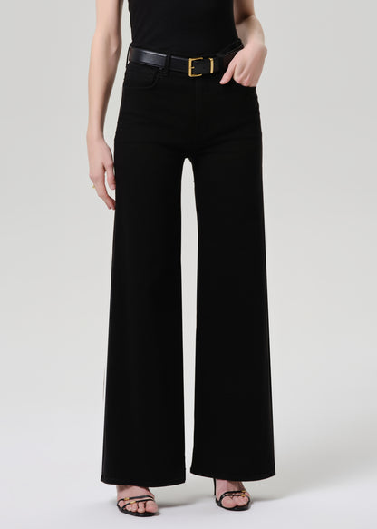 A model wearing black wide-leg trousers with a black belt featuring a gold buckle, paired with a black top and black strappy heels, against a grey background.