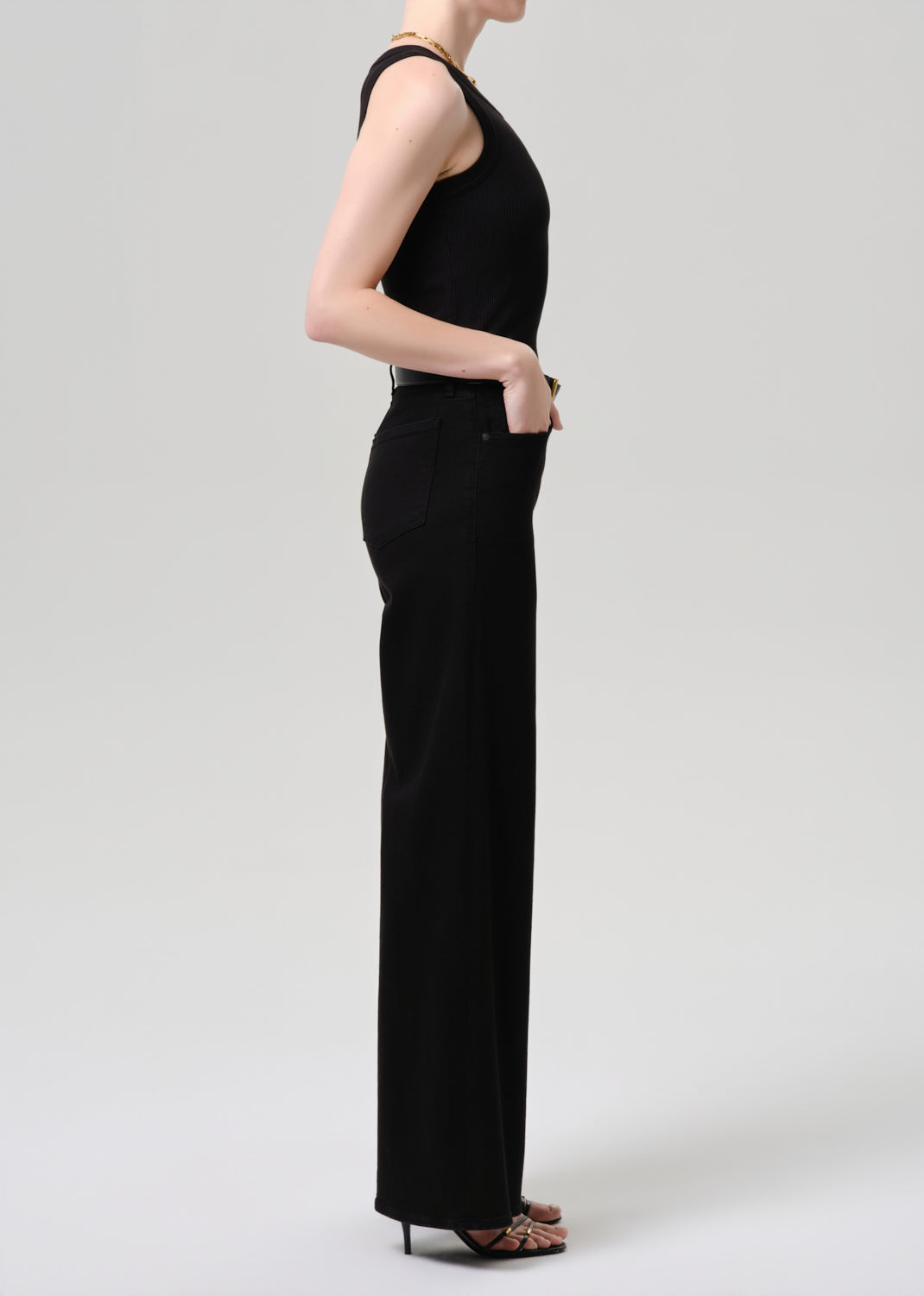 A woman shown in side profile wearing a black sleeveless high-neck jumpsuit with wide legs, paired with black high heels, against a grey  background.