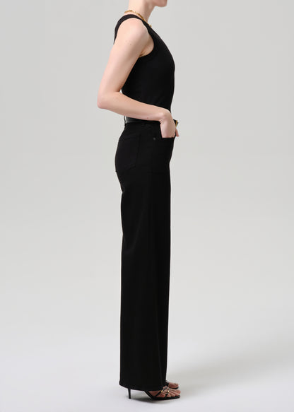 A woman shown in side profile wearing a black sleeveless high-neck jumpsuit with wide legs, paired with black high heels, against a grey  background.