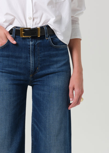 A close-up of a person wearing dark blue denim jeans with a black belt featuring a gold-tone rectangular buckle, paired with a white button-down shirt.
