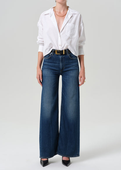 A model wearing a white button-down shirt tucked into dark blue wide-leg denim jeans with a gold belt, paired with black pointed-toe heels, against a grey background.