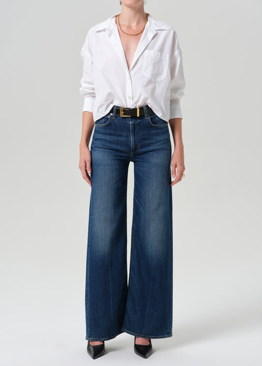 A model wearing a white button-down shirt tucked into dark blue wide-leg denim jeans with a gold belt, paired with black pointed-toe heels, against a grey background.