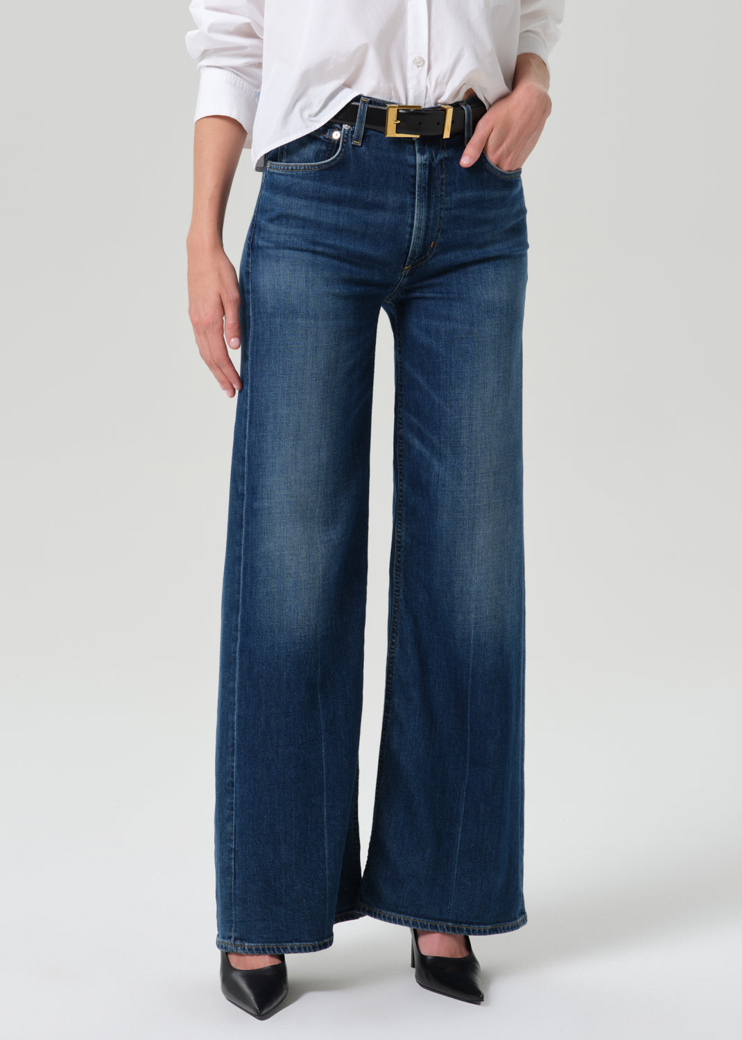A model wearing a pair of dark blue wide-leg denim jeans with a black belt featuring a gold buckle, styled with a white button-down shirt and black pointed-toe heels against a grey background.