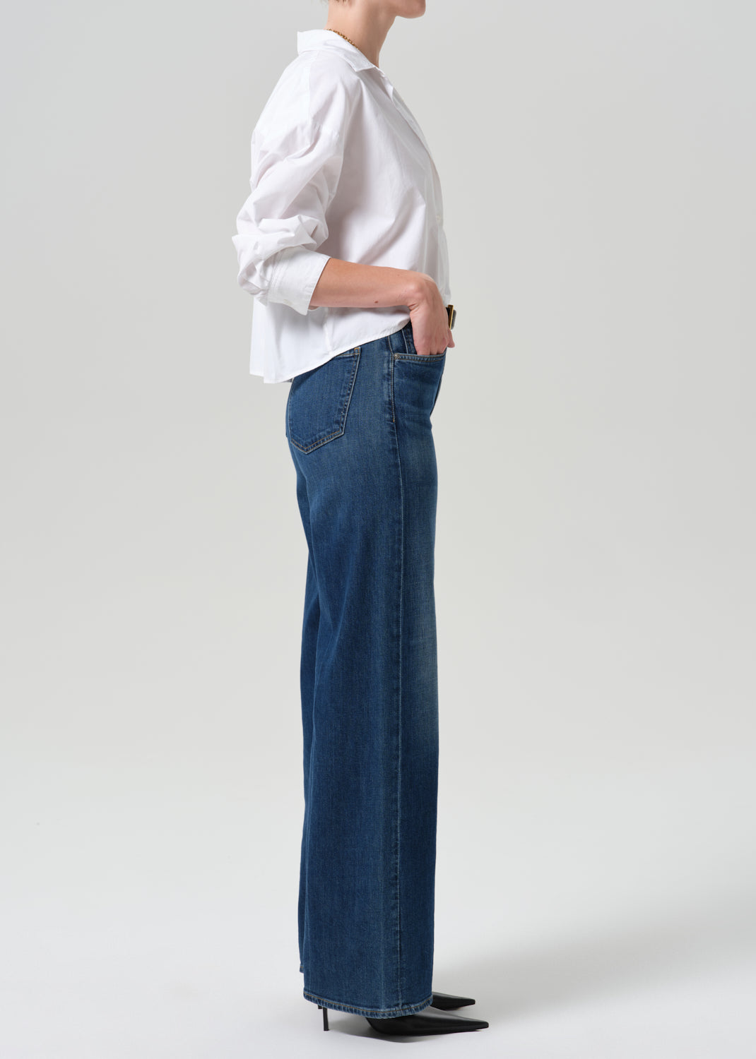 A model shown from the side wearing a white button-up shirt and dark blue wide-leg denim jeans, paired with black pointed-toe heels, against a grey background.