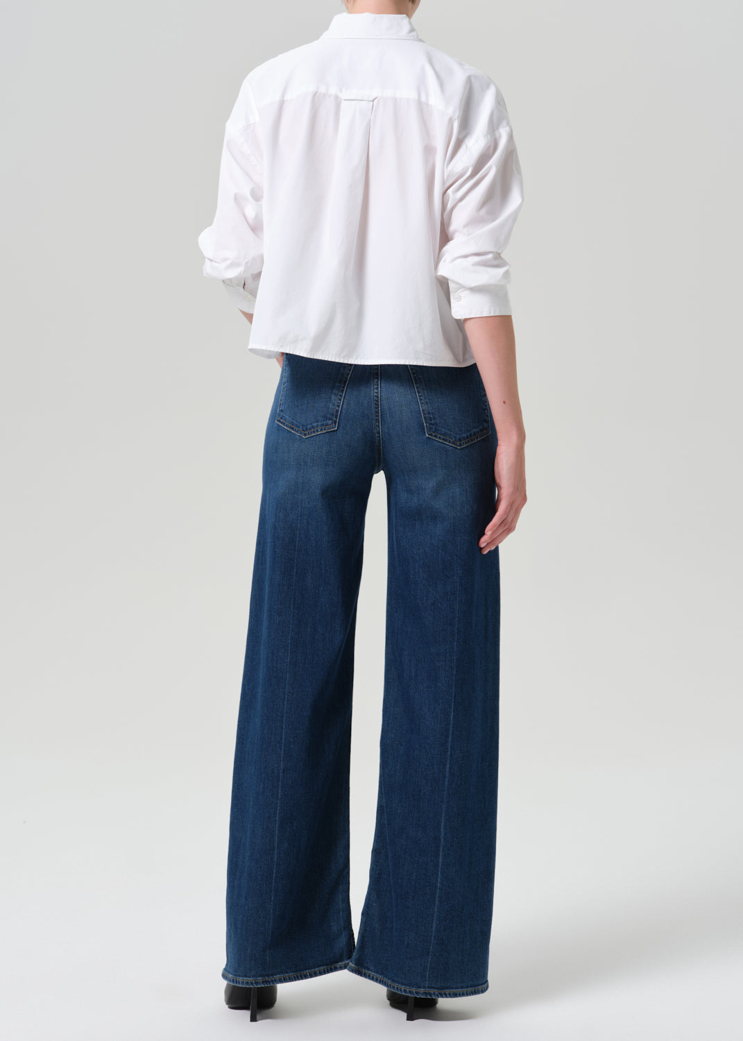 A model shown from the back wearing a white long-sleeve button-up shirt and dark blue wide-leg denim jeans, paired with black heels, against a grey background.
