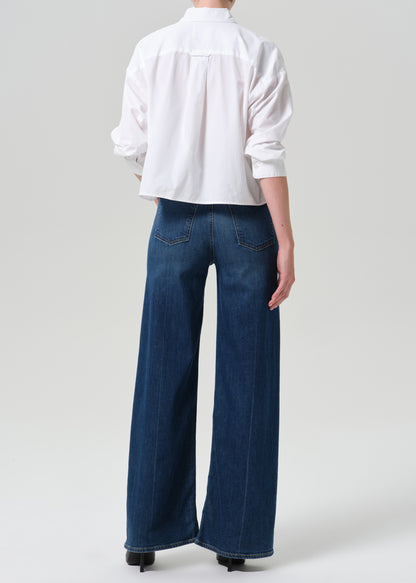 A model shown from the back wearing a white long-sleeve button-up shirt and dark blue wide-leg denim jeans, paired with black heels, against a grey background.