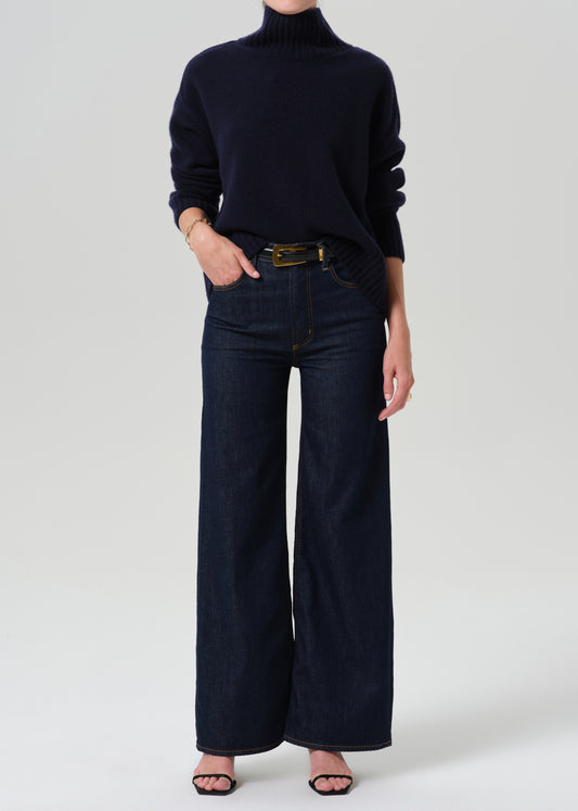 Person wearing a blue turtleneck with dark blue pants on a grey background