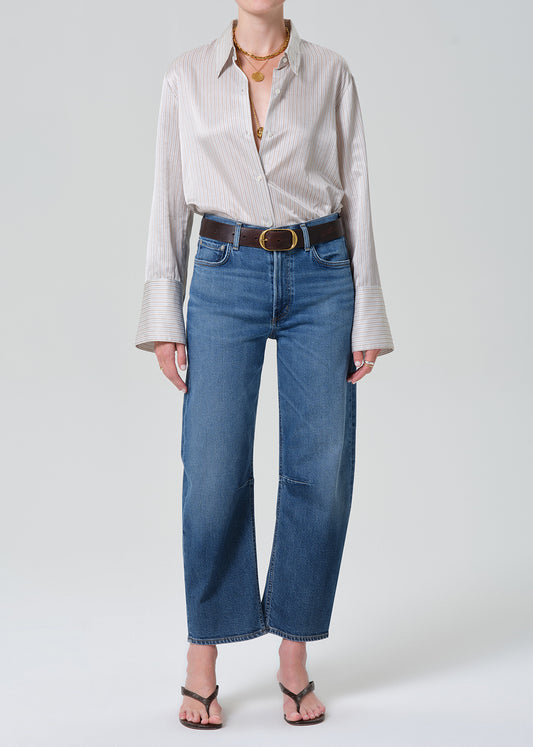 A model wearing a light beige and white vertically striped button-down shirt, blue cropped wide-leg jeans with a dark brown belt, and brown thong sandals, photographed against a grey background.