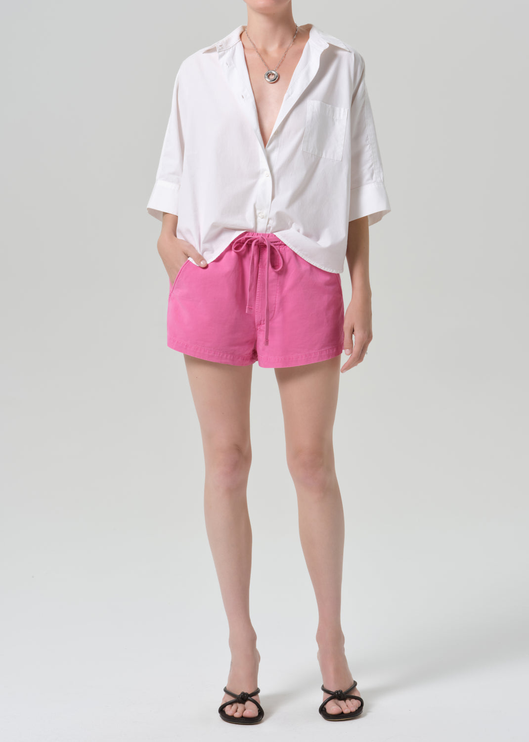 A model wearing a white short-sleeve button-up shirt tied loosely at the waist and bright pink drawstring shorts, paired with black flat sandals, against a grey background.