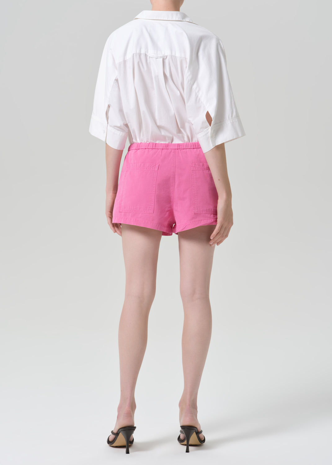 A model shown from the back wearing a white short-sleeve shirt with side cutouts and bright pink shorts, paired with black heeled sandals, against a grey background.