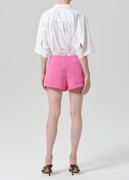 A model shown from the back wearing a white short-sleeve shirt with side cutouts and bright pink shorts, paired with black heeled sandals, against a grey background.