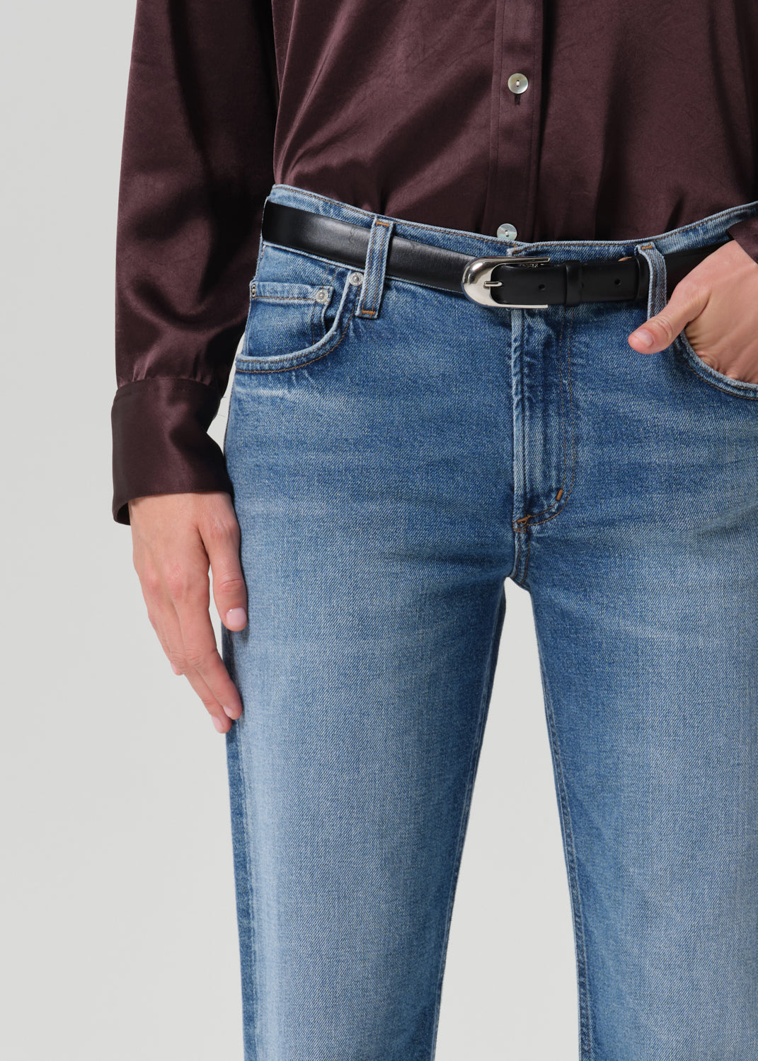 Close-up of a person wearing light blue denim jeans with a black belt and a dark burgundy button-up shirt, shown from the chest down against a grey background.