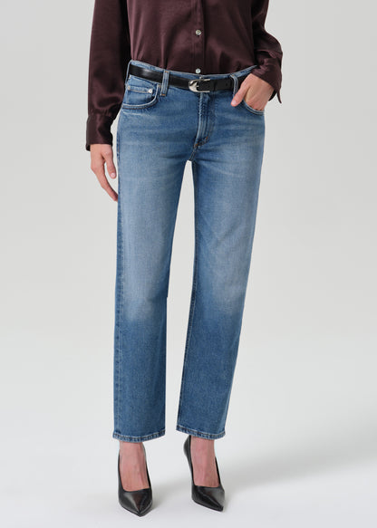 A model wearing a pair of medium-wash blue straight-leg jeans, styled with a black belt, a dark brown satin blouse, and black pointed-toe heels against a grey background.