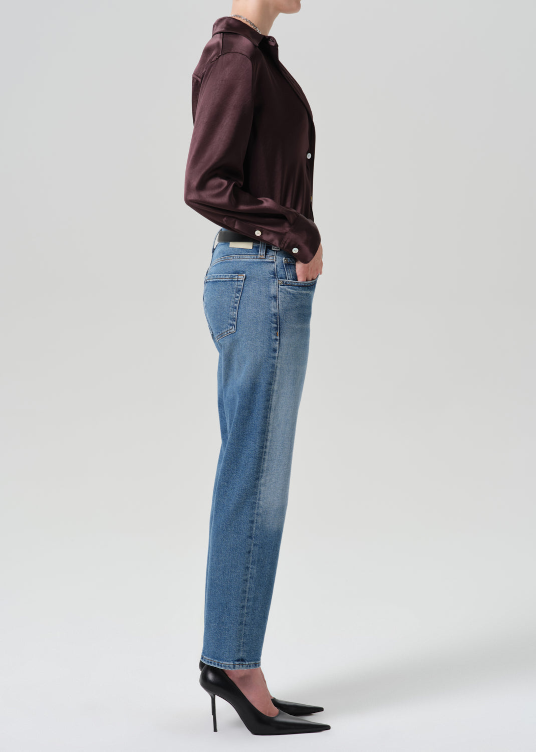 A side-profile view of a model wearing high-waisted, straight-leg blue jeans, a dark plum button-up shirt, and black pointed-toe heels against a grey background.