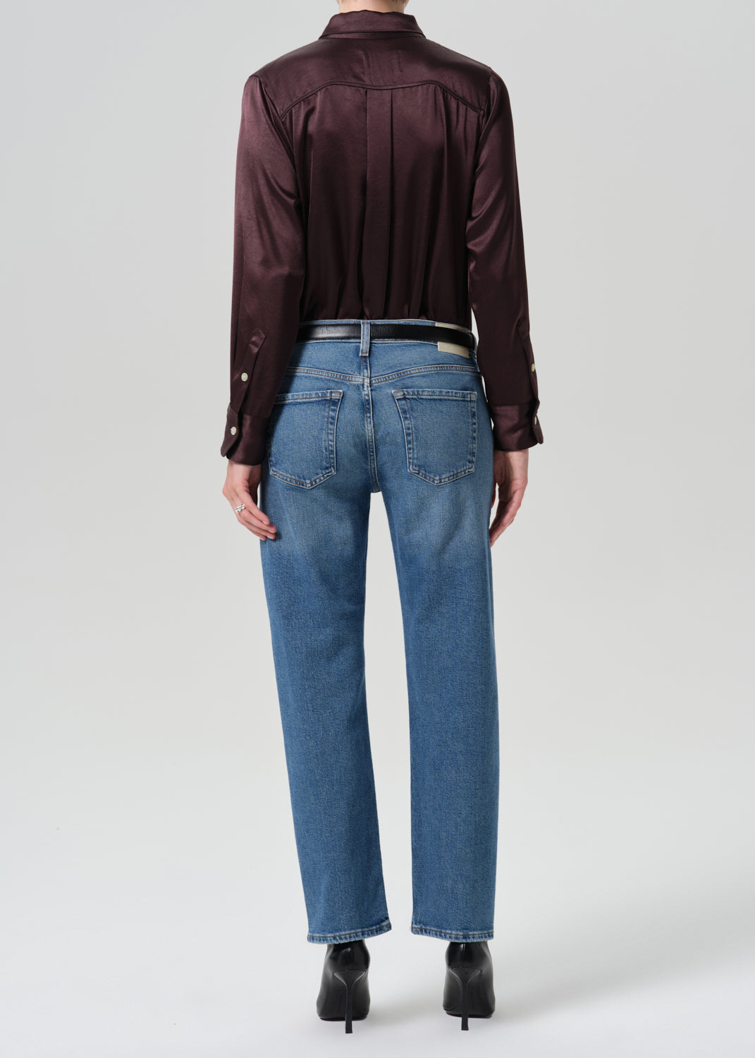 Rear view of a model wearing a dark burgundy satin shirt and blue jeans with black boots, set against a grey background.