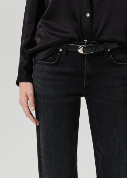 Close-up of a person wearing black jeans with a black belt featuring a silver buckle, paired with a black satin shirt, against a grey background.
