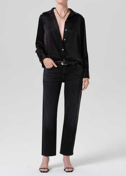 A model wearing a black long-sleeve satin button-up shirt, black jeans, a black belt, and black sandals, photographed against a grey background.