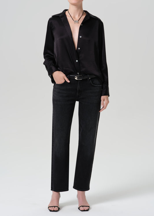 A model wearing a black long-sleeve satin button-up shirt, black jeans, a black belt, and black sandals, photographed against a grey background.