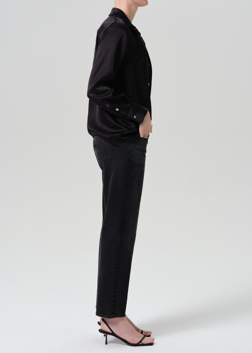 A side-profile studio shot of a model wearing a black satin button-up shirt, black jeans, and black strappy high-heeled sandals against a grey background.