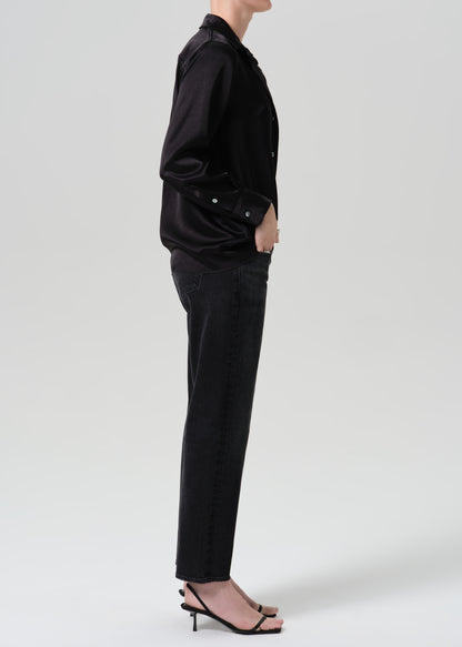 A side-profile studio shot of a model wearing a black satin button-up shirt, black jeans, and black strappy high-heeled sandals against a grey background.