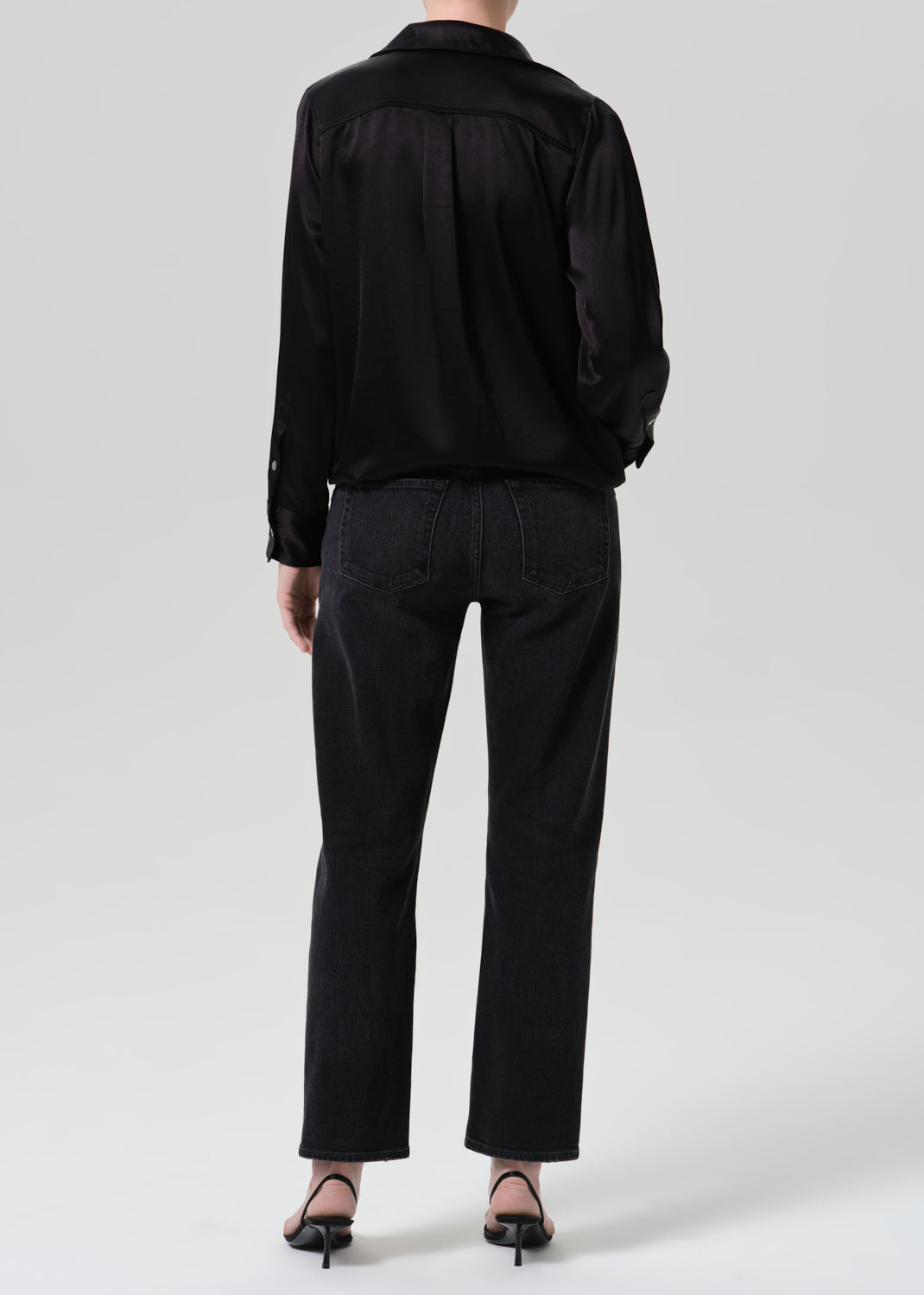 A model shown from the back wearing a black satin long-sleeve shirt and black wide-leg jeans, paired with black pointed-toe heels, against a grey background.