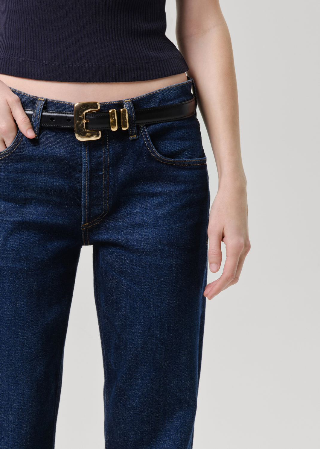 Close-up of a person wearing dark blue denim jeans with a black leather belt featuring a prominent gold-tone buckle, paired with a dark ribbed top against a grey background.