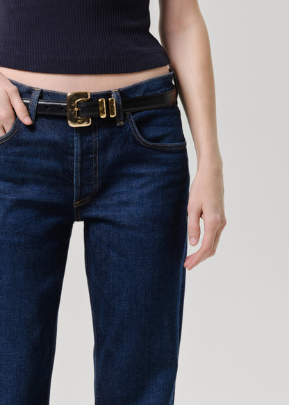 Close-up of a person wearing dark blue denim jeans with a black leather belt featuring a prominent gold-tone buckle, paired with a dark ribbed top against a grey background.