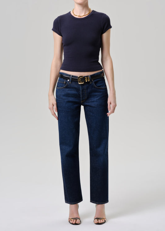 A model wearing dark blue straight-leg jeans with a gold-tone belt, paired with a navy blue short-sleeve top and black sandals, photographed against a grey background.