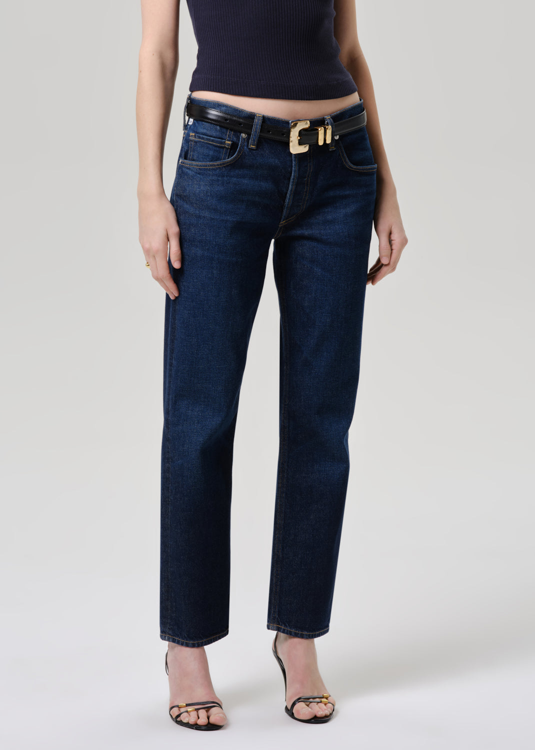 A model wearing dark blue straight-leg jeans with a black leather belt featuring a gold-tone buckle, paired with a navy top and black strappy heels, against a grey background.