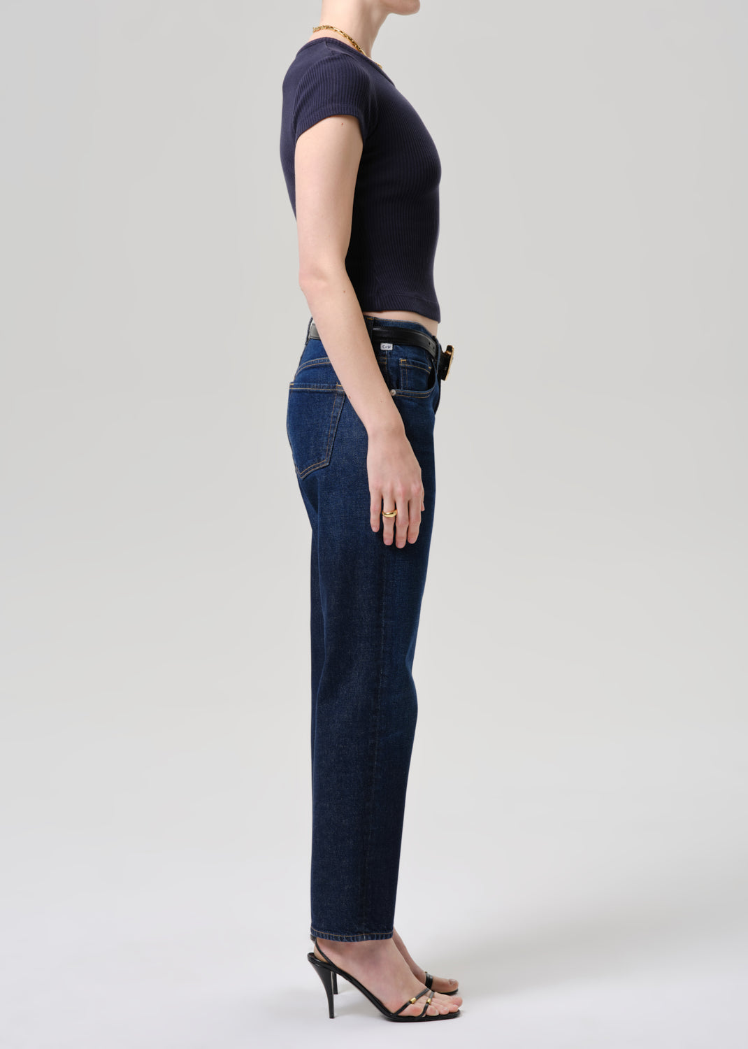 A side-profile studio shot of a model wearing dark blue straight-leg jeans, a dark navy short-sleeve crop top, and black heeled sandals against a grey background.