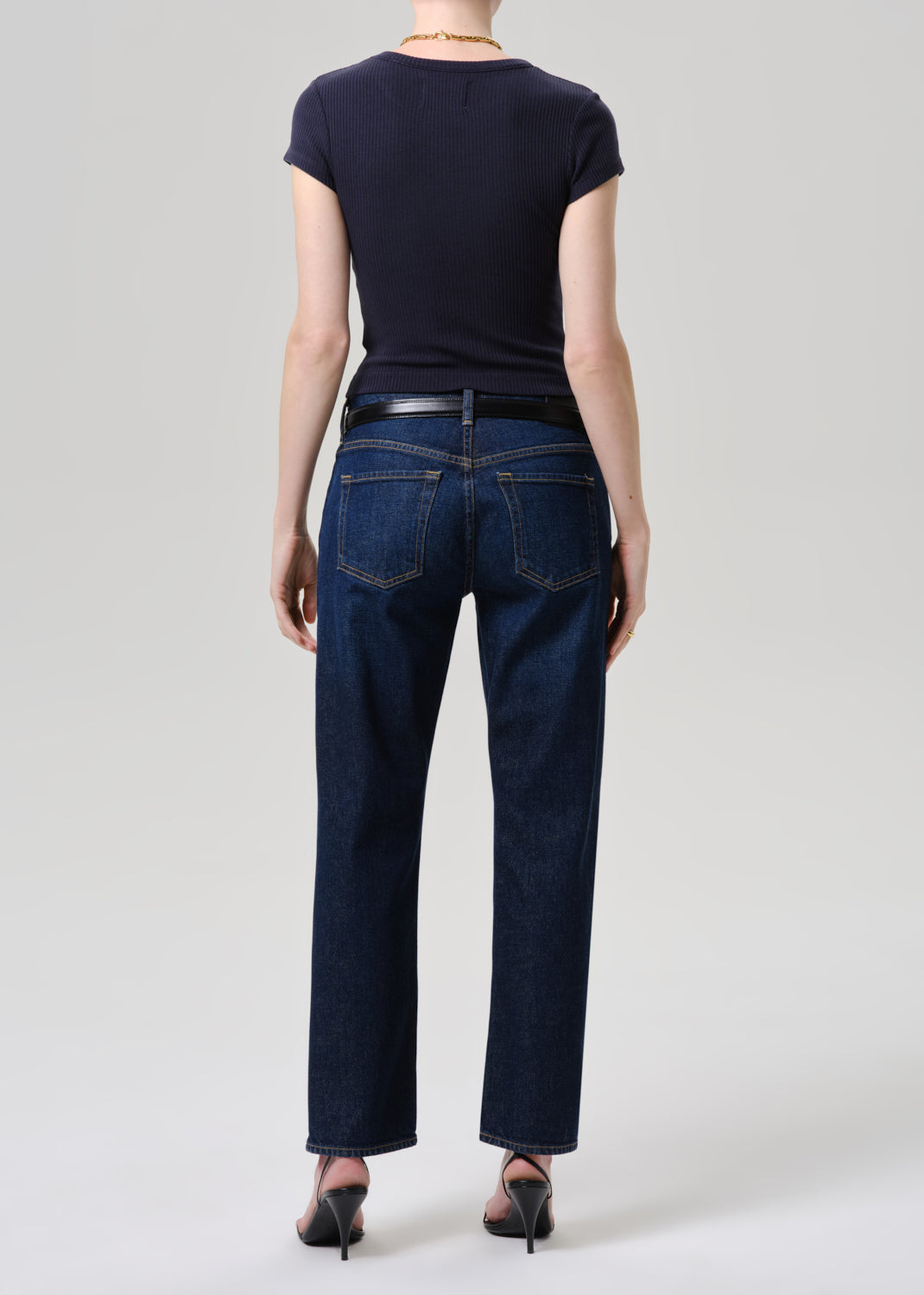 A model shown from the back wearing dark blue straight-leg jeans, a black short-sleeve top, a black belt, and black pointed-toe heels against a grey background.