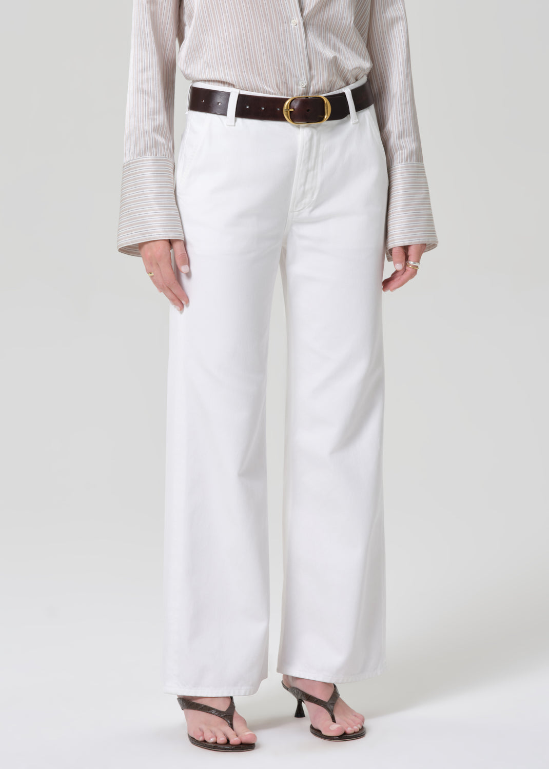 Person wearing a light-colored shirt and white jeans with a black belt on a plain background