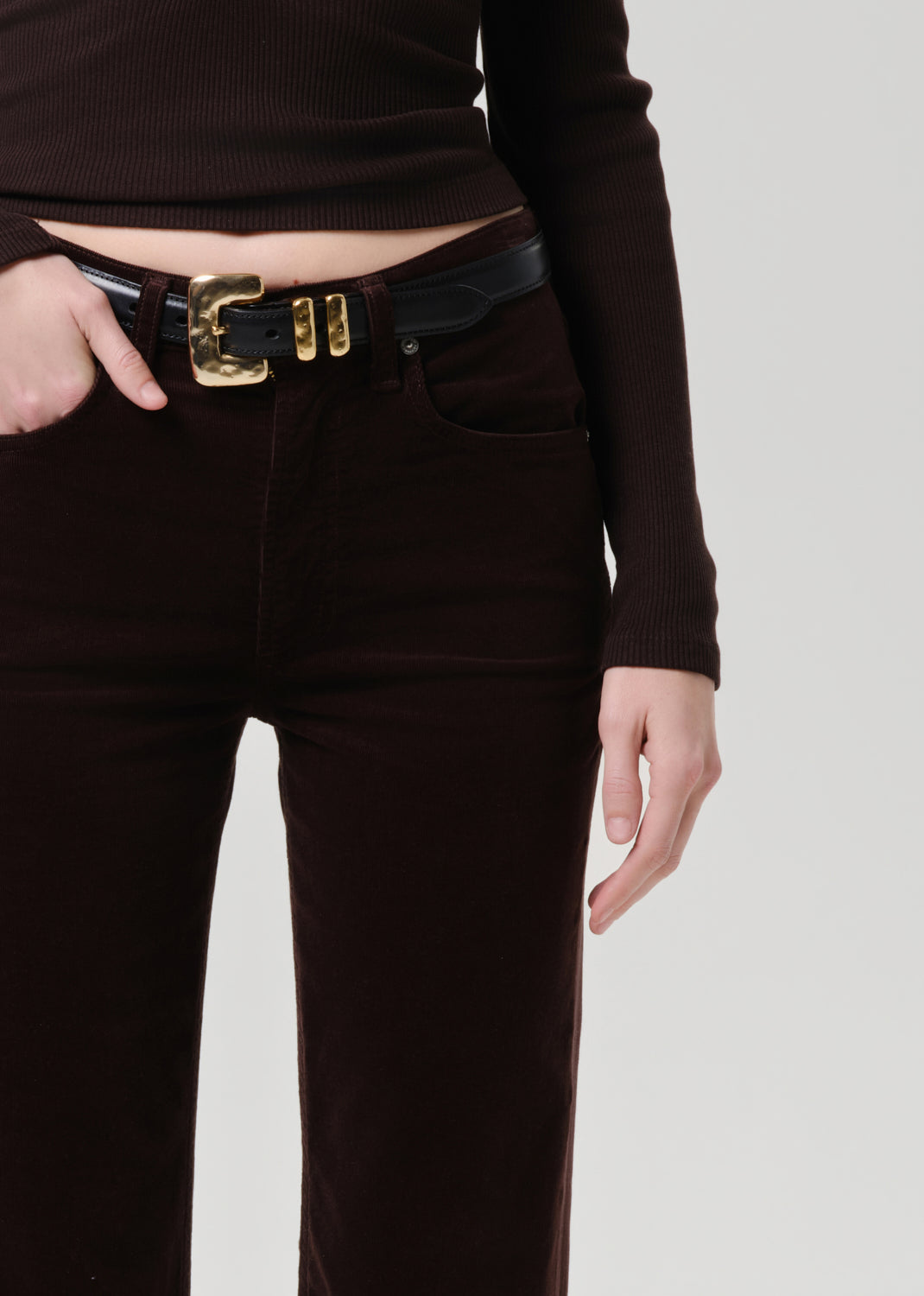 Person wearing brown pants with a black belt and gold buckle on a grey background