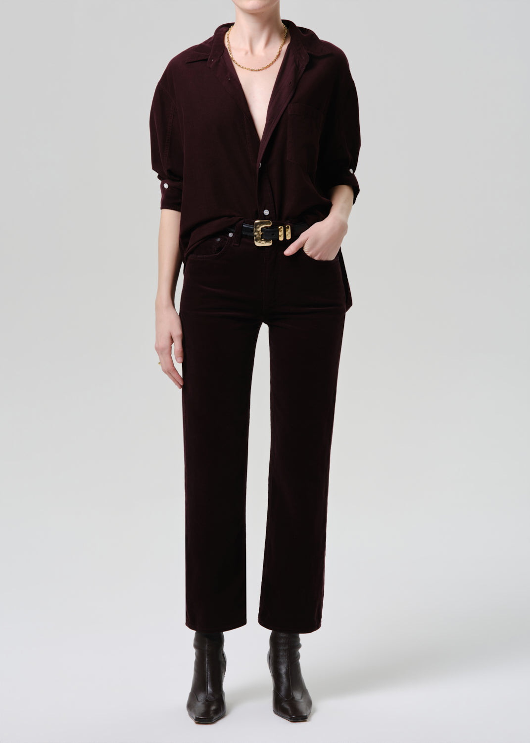 Person wearing a dark brown outfit with a belt and boots on a grey background