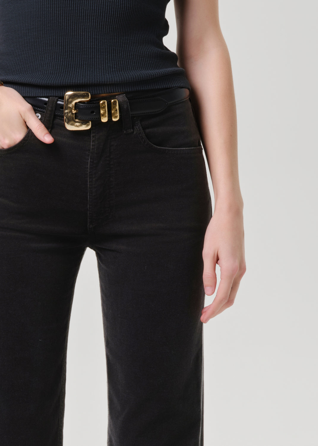 Close-up of black straight leg jeans showing the waistband, front pockets, and black belt with gold buckle.