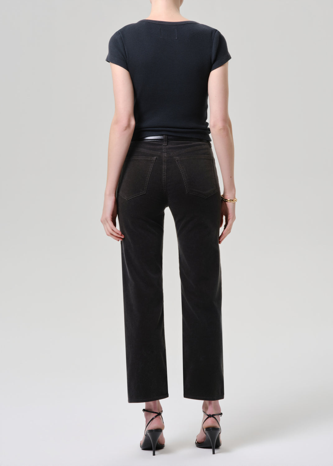 Person wearing black straight leg cropped jeans with a black short sleeve fitted top and black heels, back full body view on a grey background.