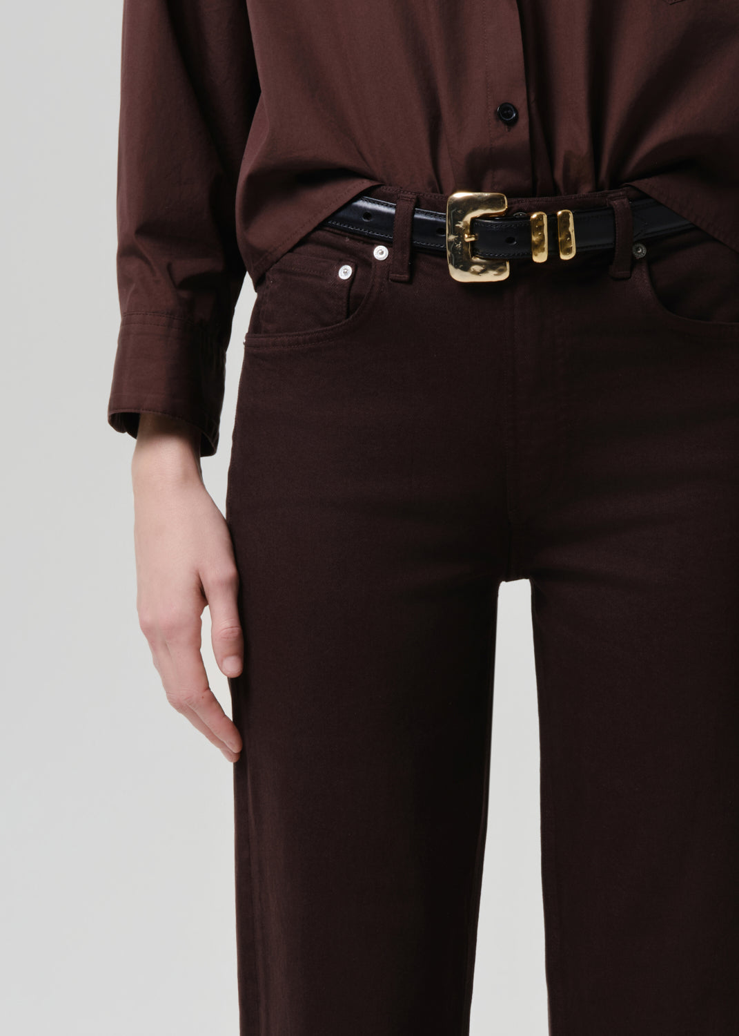 Person wearing a brown shirt and pants with a black belt on a grey background
