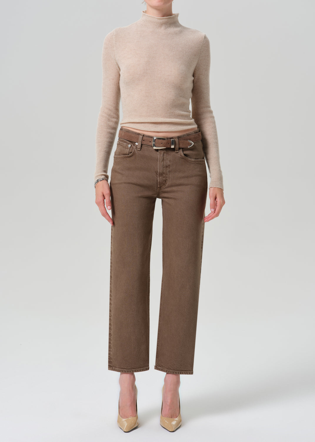 A model wearing a beige ribbed turtleneck sweater and brown wide-leg cropped jeans with a matching belt, paired with beige heels, against a grey background.