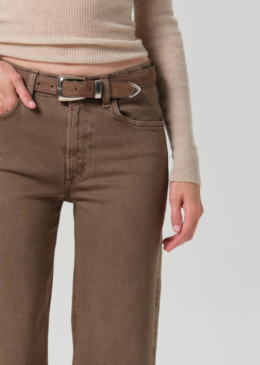 Close-up of a person wearing brown jeans with a matching brown belt featuring a silver-toned buckle, paired with a beige ribbed knit top against a grey background.